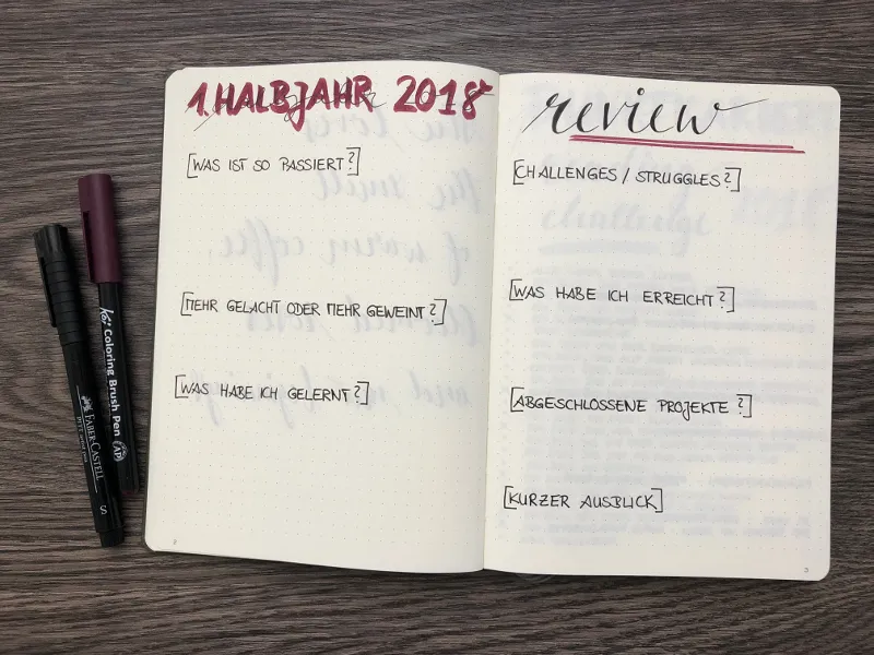 Aufgeschlagenes Notizbuch mit einer Doppelseite zum Rückblick, oben die Überschriften 1. Halbjahr 2018 und Review, darunter Fragen in eckigen Klammern wie Was habe ich gelernt und Kurzer Ausblick, daneben 2 Stifte