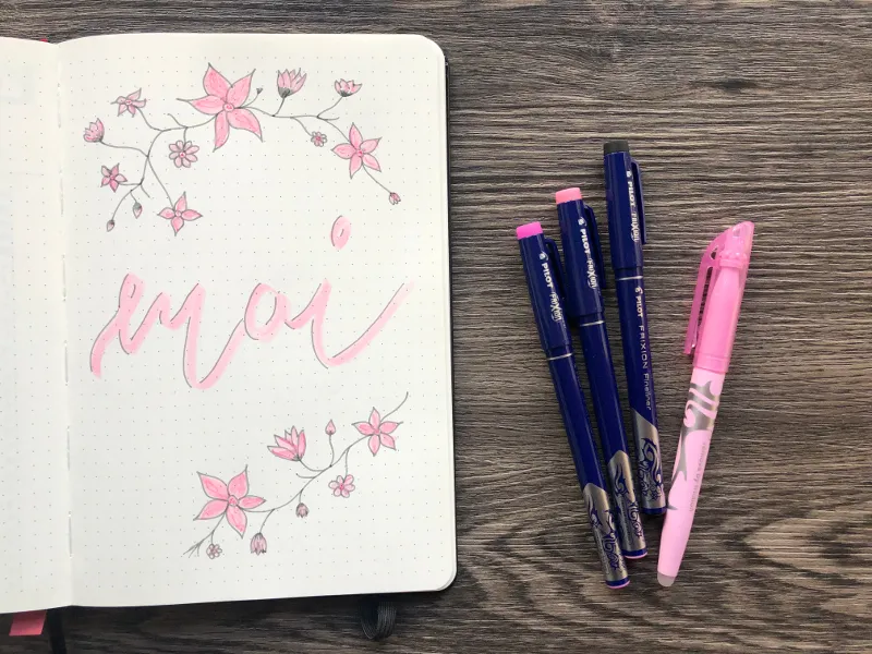 Bullet-Journal-Deckblatt mit rosa Kirschblueten und Mai-Brushlettering neben Pilot-Stiften auf Holztisch