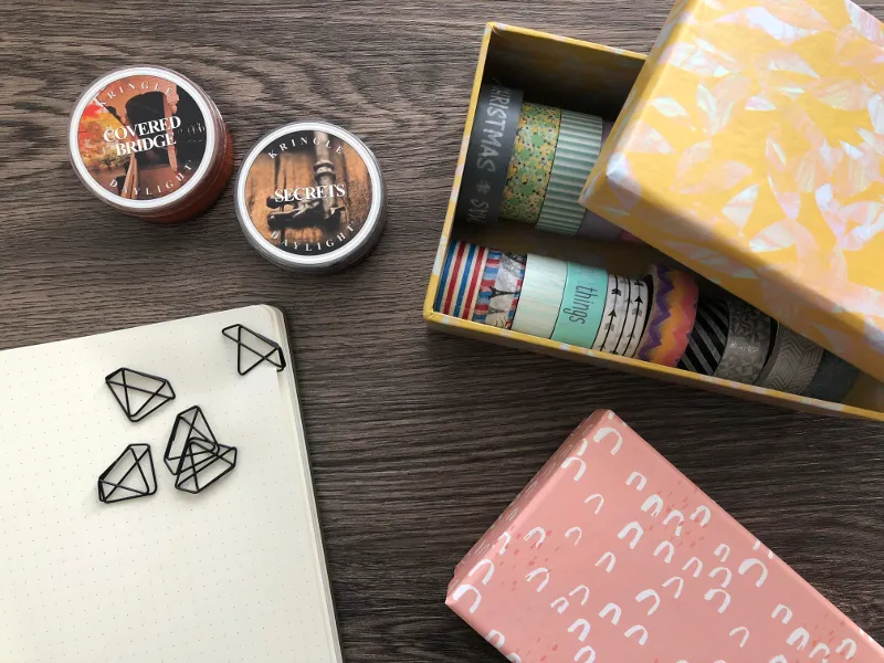 Washi-Tape-Boxen, zwei Kerzen und Büroklammern in Diamantform neben Notizbuch