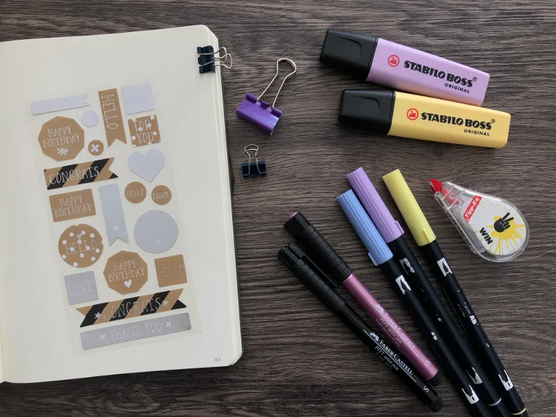 Stickerseite mit Stabilo-Textmarkern, Fineliner und Brush-Pens auf Holztisch