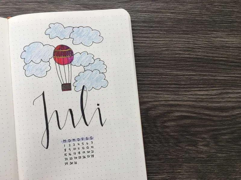Bullet-Journal-Deckblatt für Juli mit gemaltem bunten Heißluftballon, blauen Wolken und Mini-Kalender