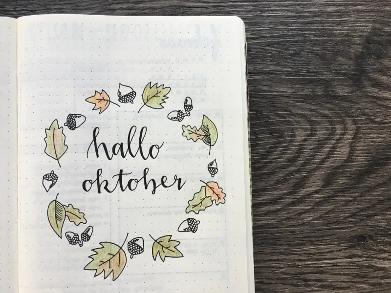 Deckblatt mit dem Schriftzug hallo oktober in einem Kranz aus kolorierten Herbstblättern und Eicheln