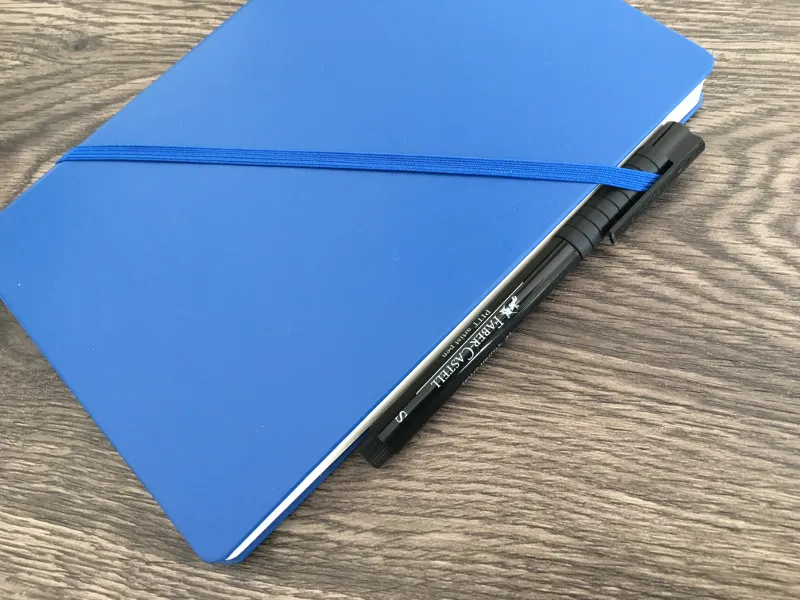 Blaues Notizbuch mit schwarzem Stift unter dem diagonalen Gummiband als Stiftschlaufe