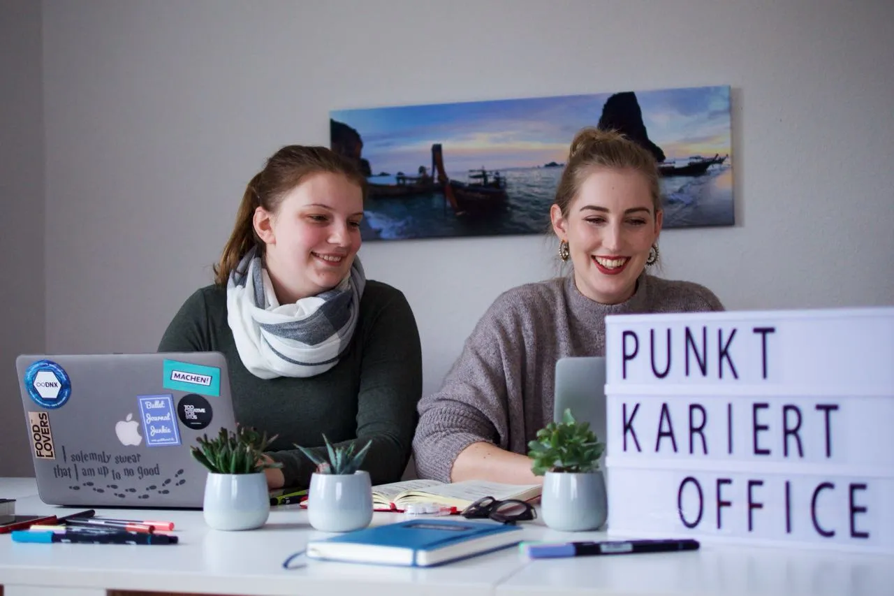 Zwei Frauen lachen am Schreibtisch neben Leuchtbox mit Schriftzug Punkt Kariert Office